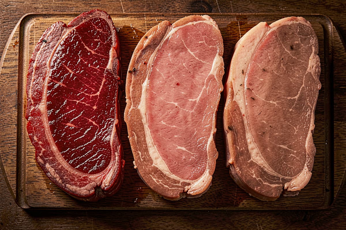 Cross-section of three steaks at different doneness levels — rare, medium-rare, medium — on a wooden cutting board