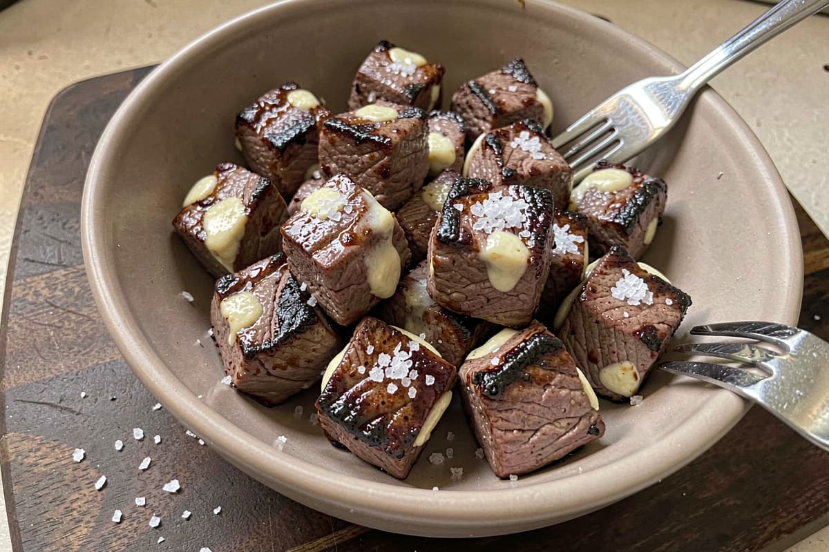 Bowl of air fryer steak bites, cube-shaped pieces with deep brown sear on all sides, glossy with butter, scattered salt flakes, fork resting on the rim