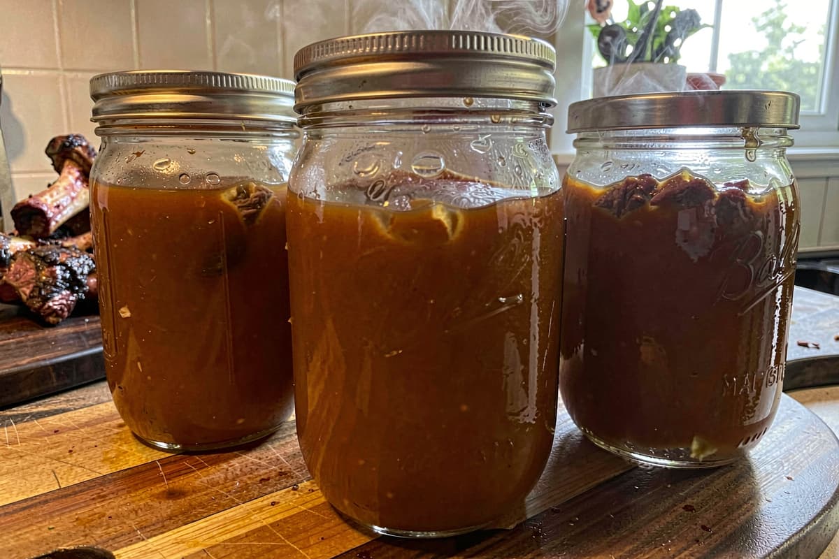 Glass mason jars of homemade beef broth on a wooden counter, deep amber color, gelatinous when cold, with roasted beef bones visible in the background