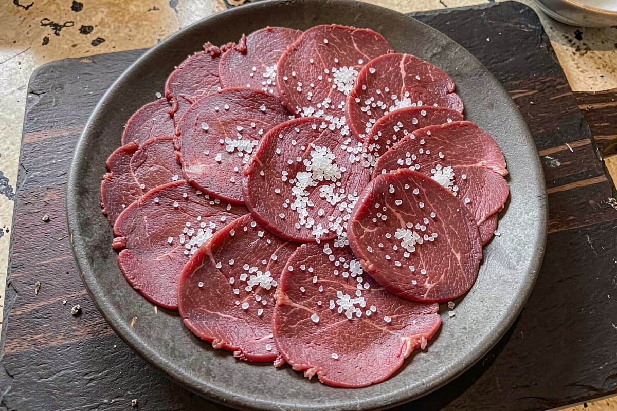 Beef carpaccio on a chilled plate, paper-thin slices of raw red beef arranged in a single overlapping layer, scattered coarse salt across the surface, dark slate background