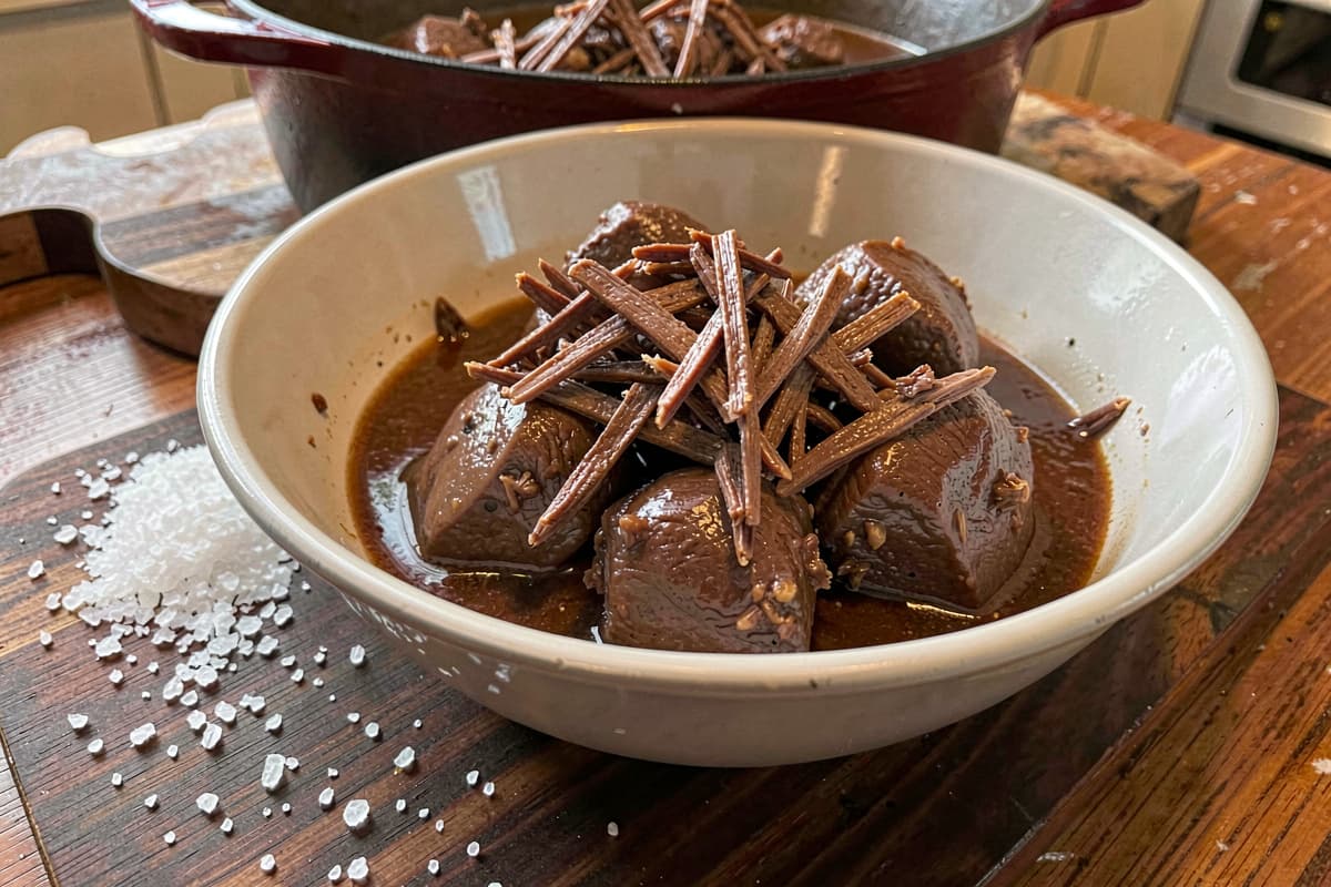 Fork-shredded braised beef cheeks in a wide bowl, deep brown meat in glossy braising liquid, Dutch oven visible in the background, scattered salt on the wooden table