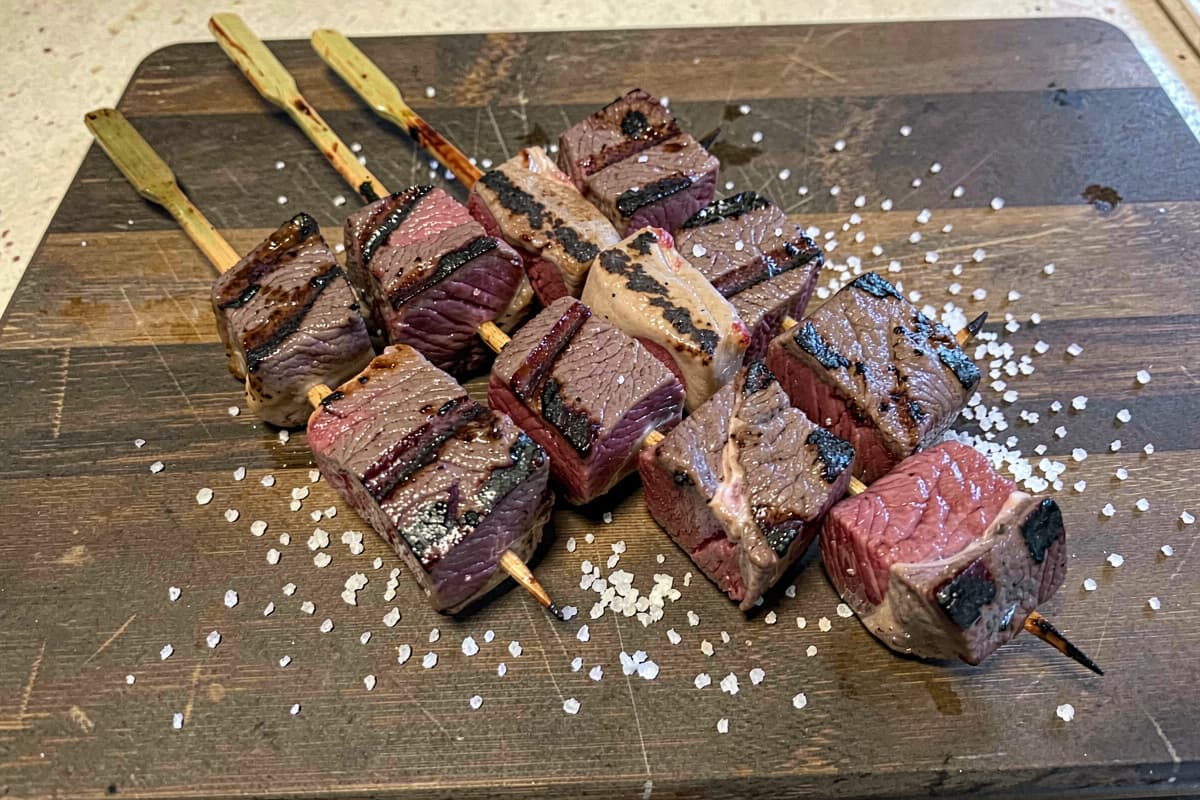 Beef heart skewers on a wooden cutting board, deep brown grill marks, dark red medium-rare interior visible at the cube edges, scattered salt, four skewers fanned out