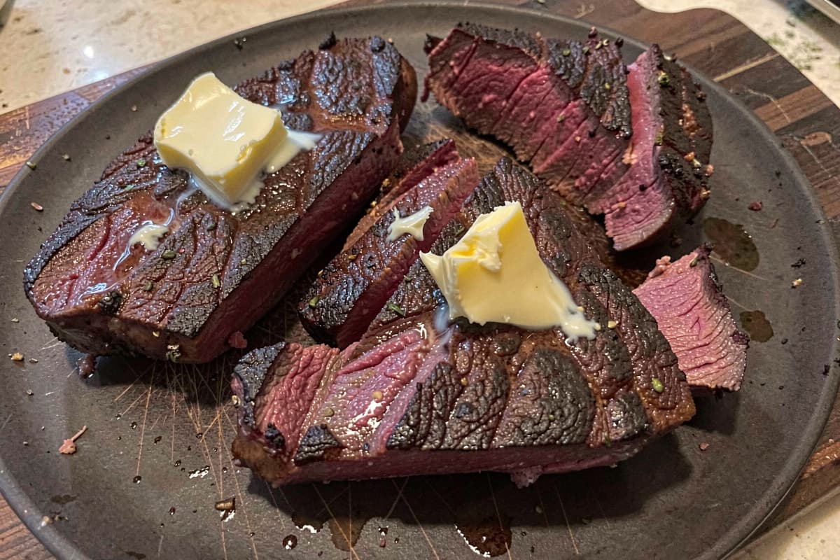 Pan-seared beef heart steaks on a wooden cutting board, deep brown crust, dark red medium-rare interior, sliced across the grain, butter glistening on top