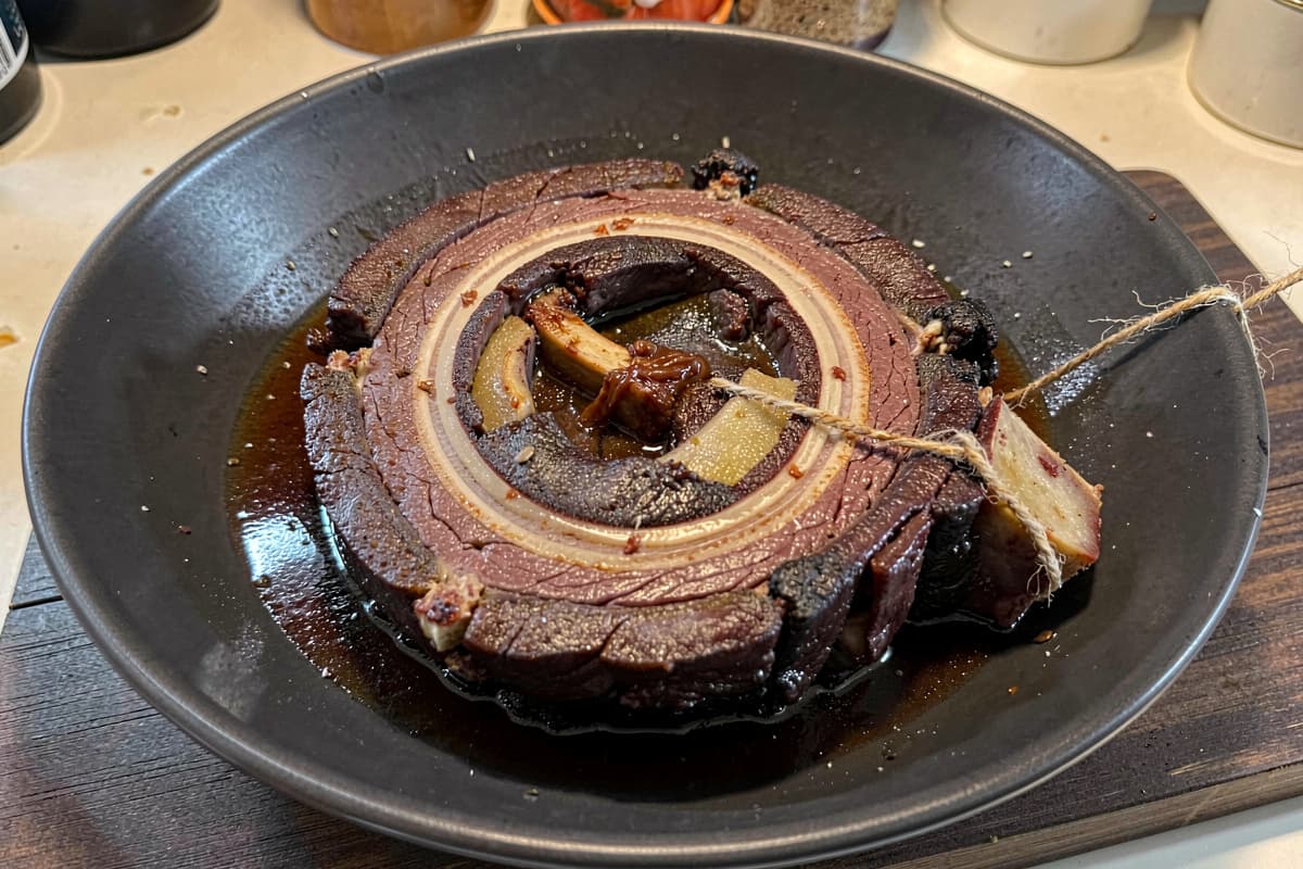 Cross-cut beef shank slab in a wide bowl with rich braising liquid, marrow visible in the center bone, deep brown braised meat ring surrounding it, twine snipped on the side