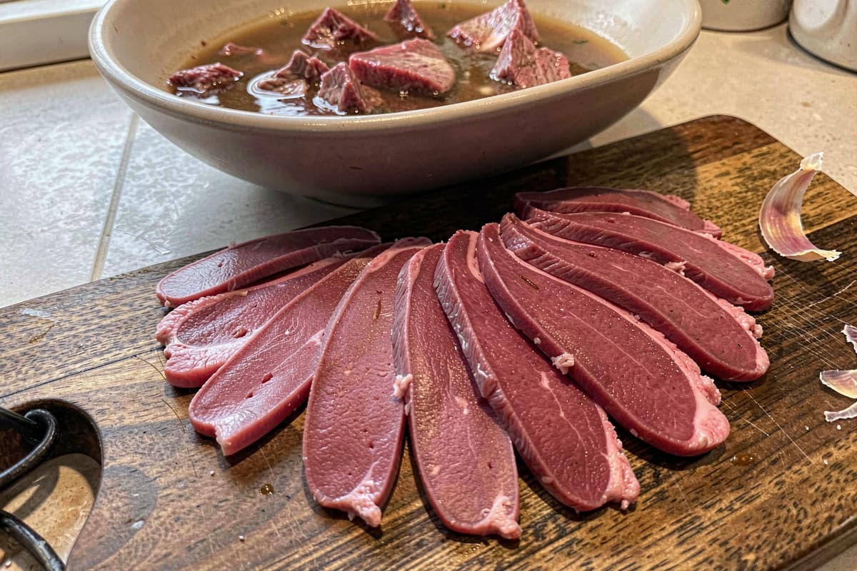 Sliced beef tongue on a wooden cutting board, deep pink interior, thin slices fanned out, peel set aside on the board, rich beef broth in a bowl behind it