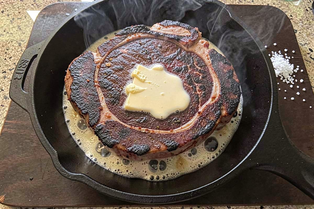 Whole ribeye steak in a cast iron skillet on a stovetop, deep brown crust on top, butter foaming around it, smoke rising slightly, salt scattered nearby