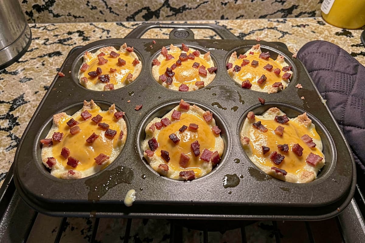Six baked carnivore egg and cheese cups with melted cheddar and crumbled bacon in a black muffin tin
