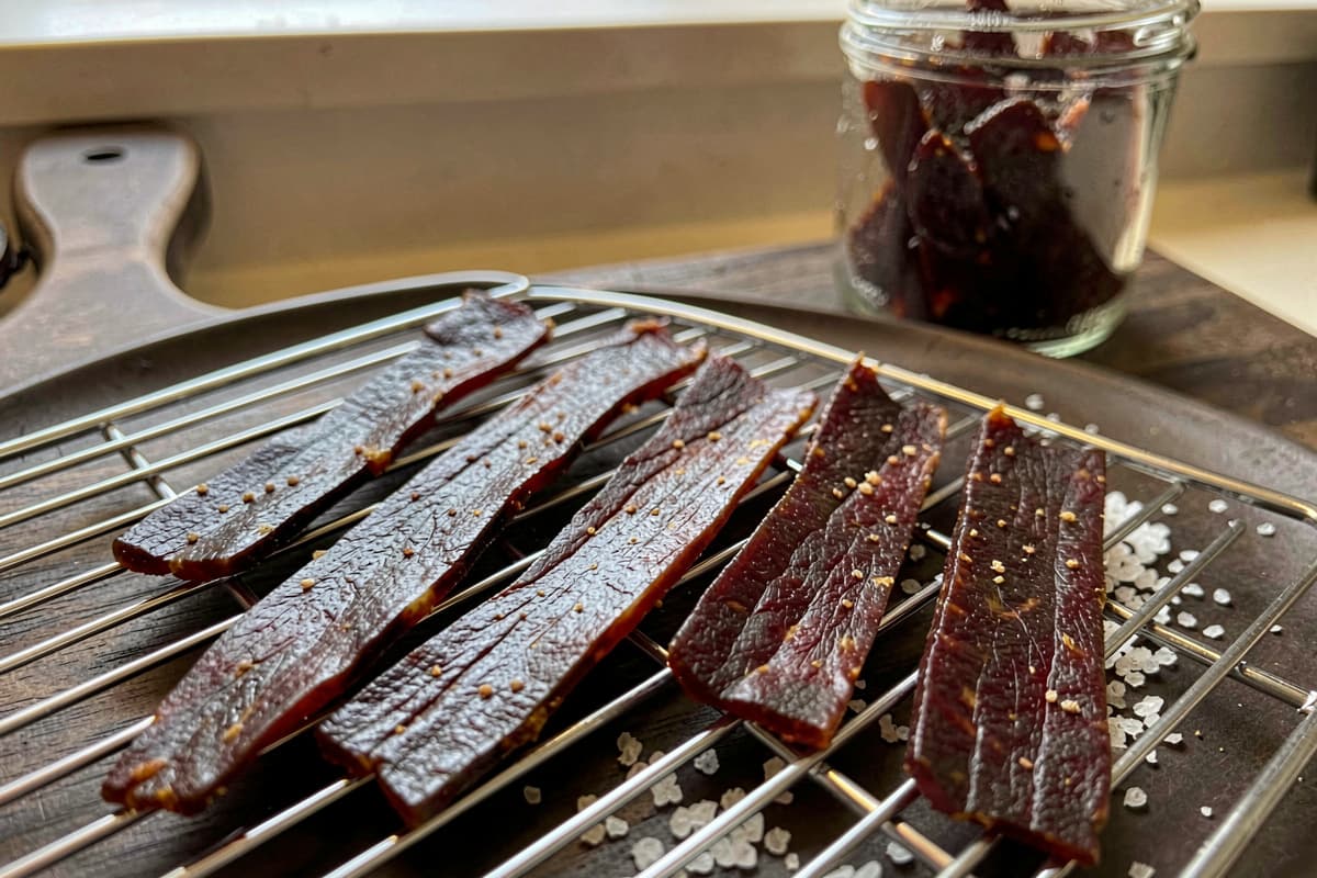 Strips of homemade beef jerky on a wire rack, dark mahogany color, slightly textured surface, scattered salt, glass jar of finished jerky in the background