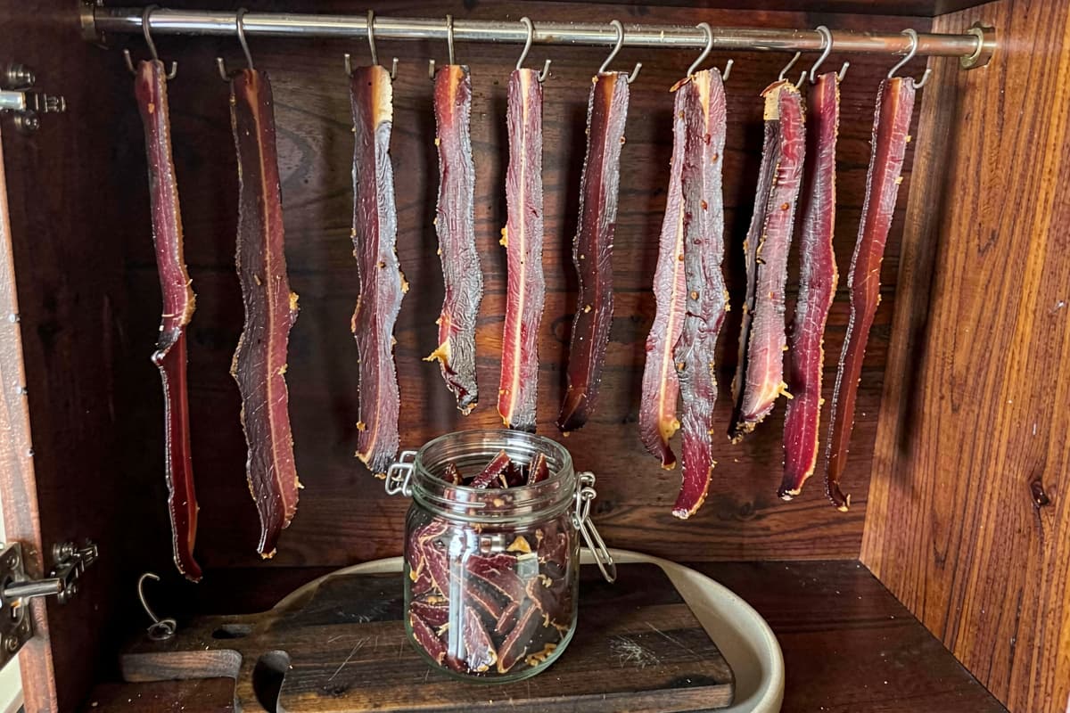 Strips of air-dried biltong hanging from butcher hooks in a wooden drying box, deep mahogany surface, varying thicknesses, glass jar of finished biltong below them