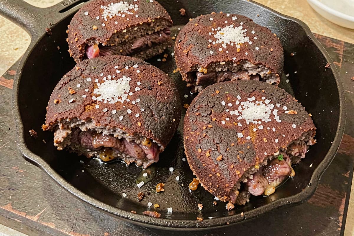 Four lamb burgers in a cast iron skillet, deep brown crusty exterior, juicy medium interior visible at the cut, scattered salt, glistening with rendered fat