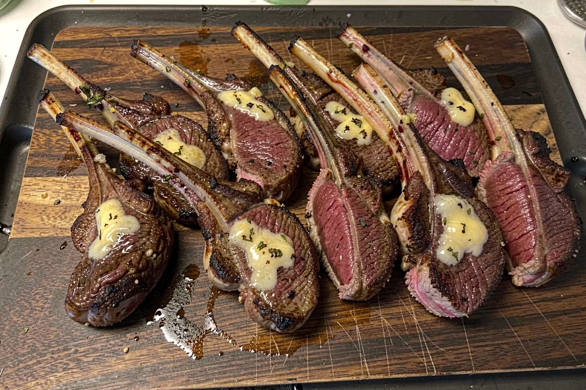 Eight pan-seared lamb loin chops on a wooden cutting board, deep brown crust, rosy medium-rare interior visible at the cut, T-shaped bones prominent, butter glistening on top