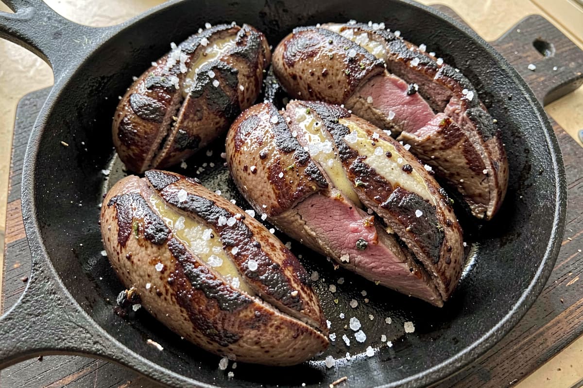 Pan-seared lamb kidneys in a cast iron skillet, split lengthwise with deep brown crusty exteriors, glossy with butter, slight pink at the cut interior, scattered salt
