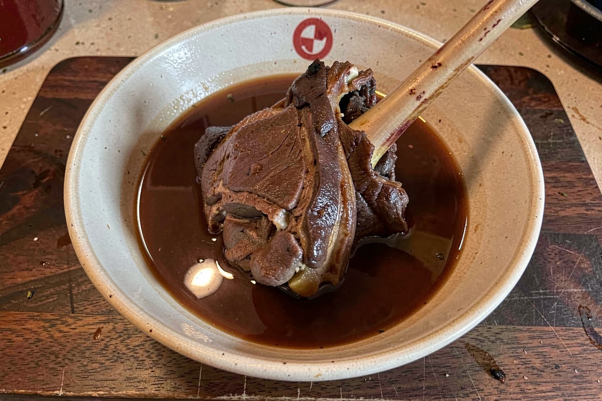 Braised lamb shank in a wide bowl with rich broth, deep brown meat falling off the bone, lamb bone protruding, glossy braising liquid pooled around it