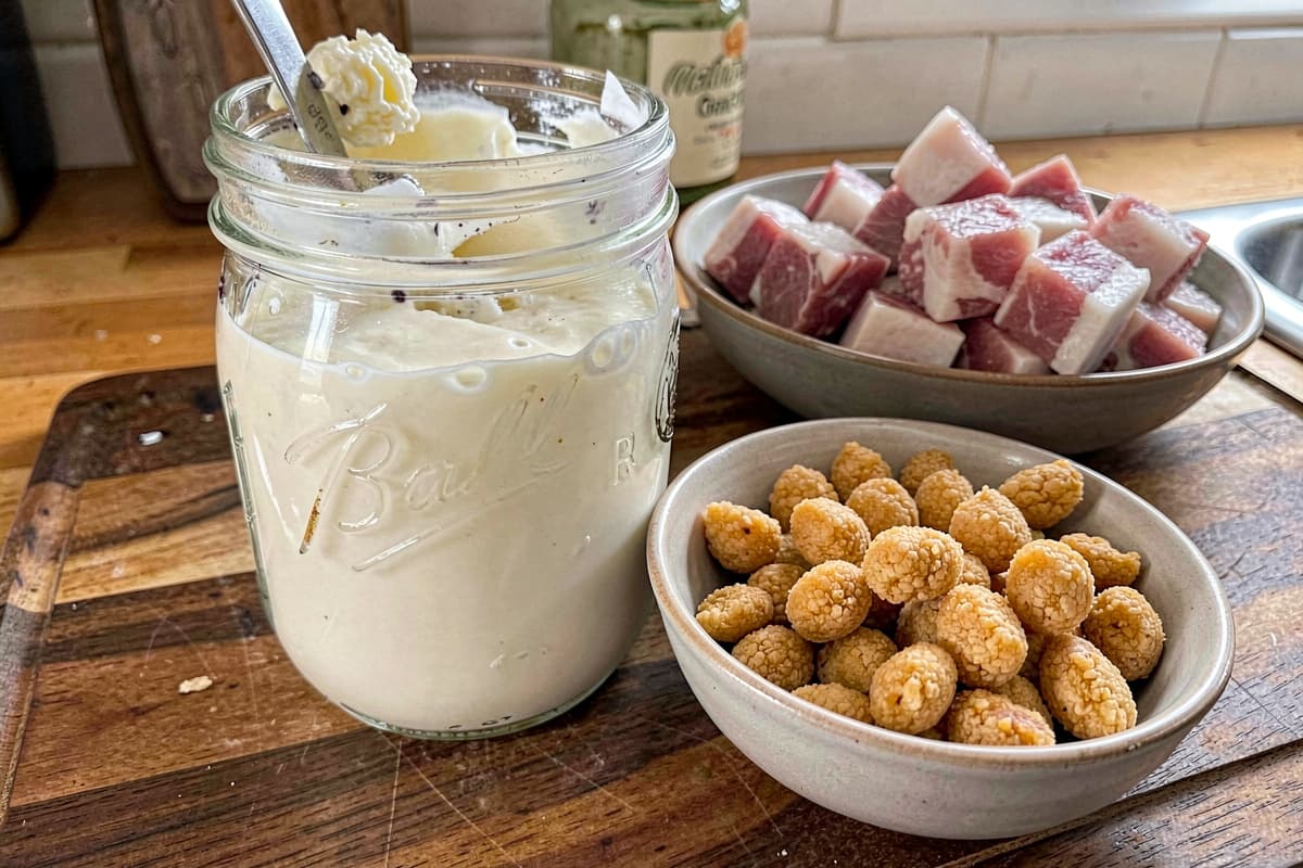 Glass mason jar of homemade lard on a wooden counter, creamy white when cooled, smaller bowl of golden cracklings beside it, raw pork fat cubes in the background