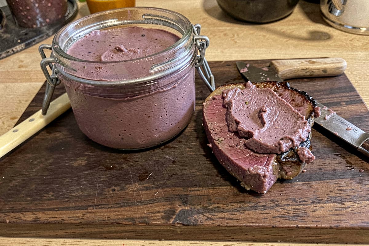 Glass jar of beef liver pâté on a wooden cutting board, smooth dark mauve surface, butter knife resting beside it, slice of cooked steak with pâté smeared on top