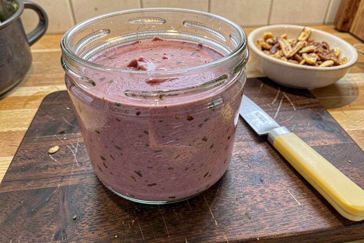 Glass jar of homemade liverwurst on a wooden cutting board, smooth pinkish-mauve surface, butter knife resting beside it, cracklings in a small bowl in the background
