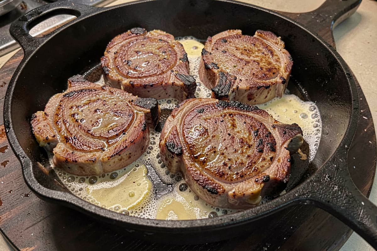 Four bone-in pork chops in a cast iron skillet just out of the oven, golden-brown crust on top, juices pooling, butter still foaming around them