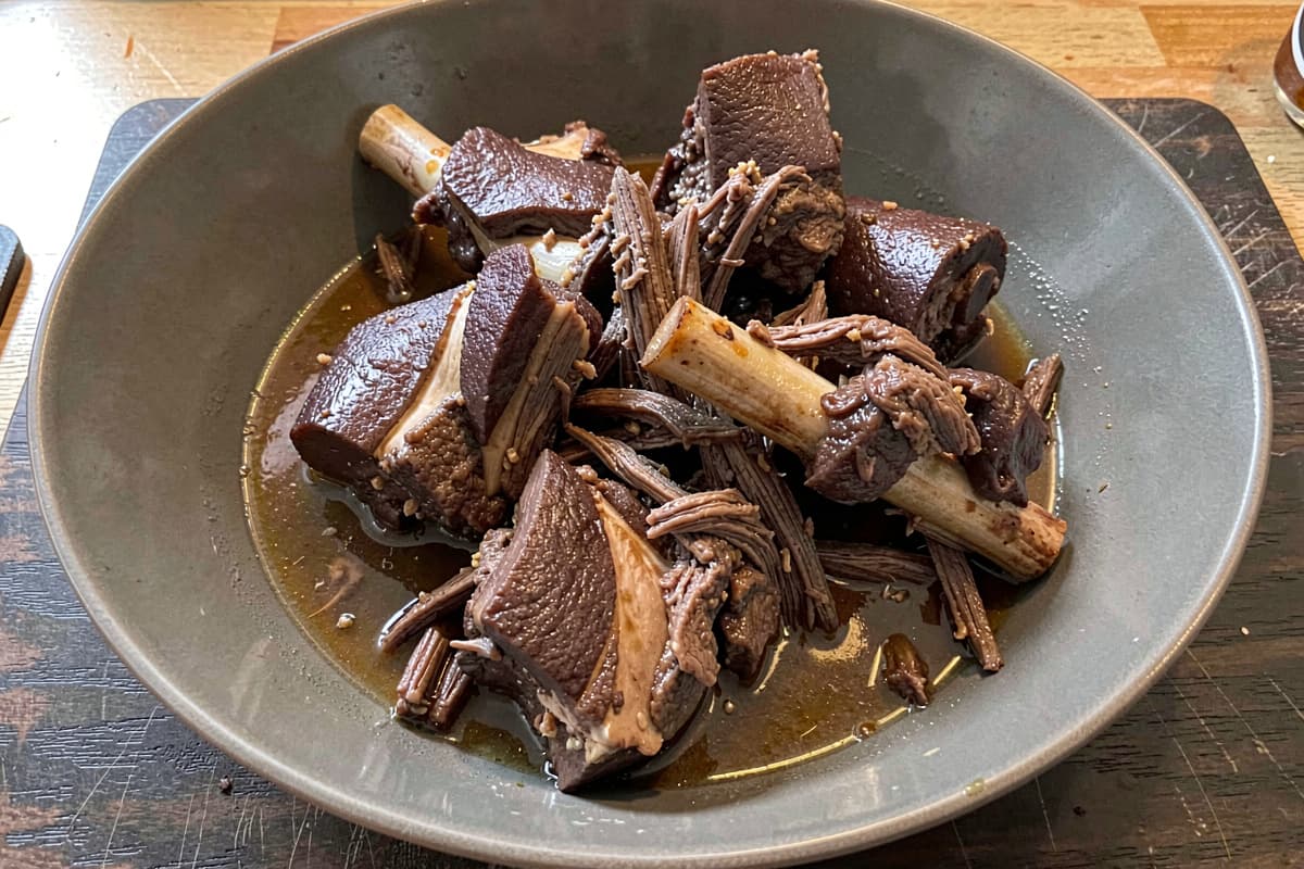 Braised oxtail segments in a wide bowl, deep brown meat falling off the bone, glossy gelatin-rich braising liquid pooled around them, shreds of meat fanning across the broth