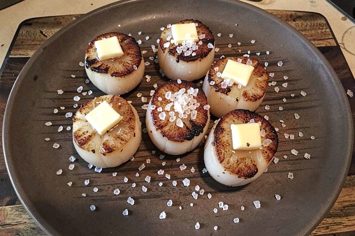 Eight pan-seared scallops on a wooden plate, deep golden-brown crust on top, scattered salt, butter glistening on each scallop, dark wood underneath
