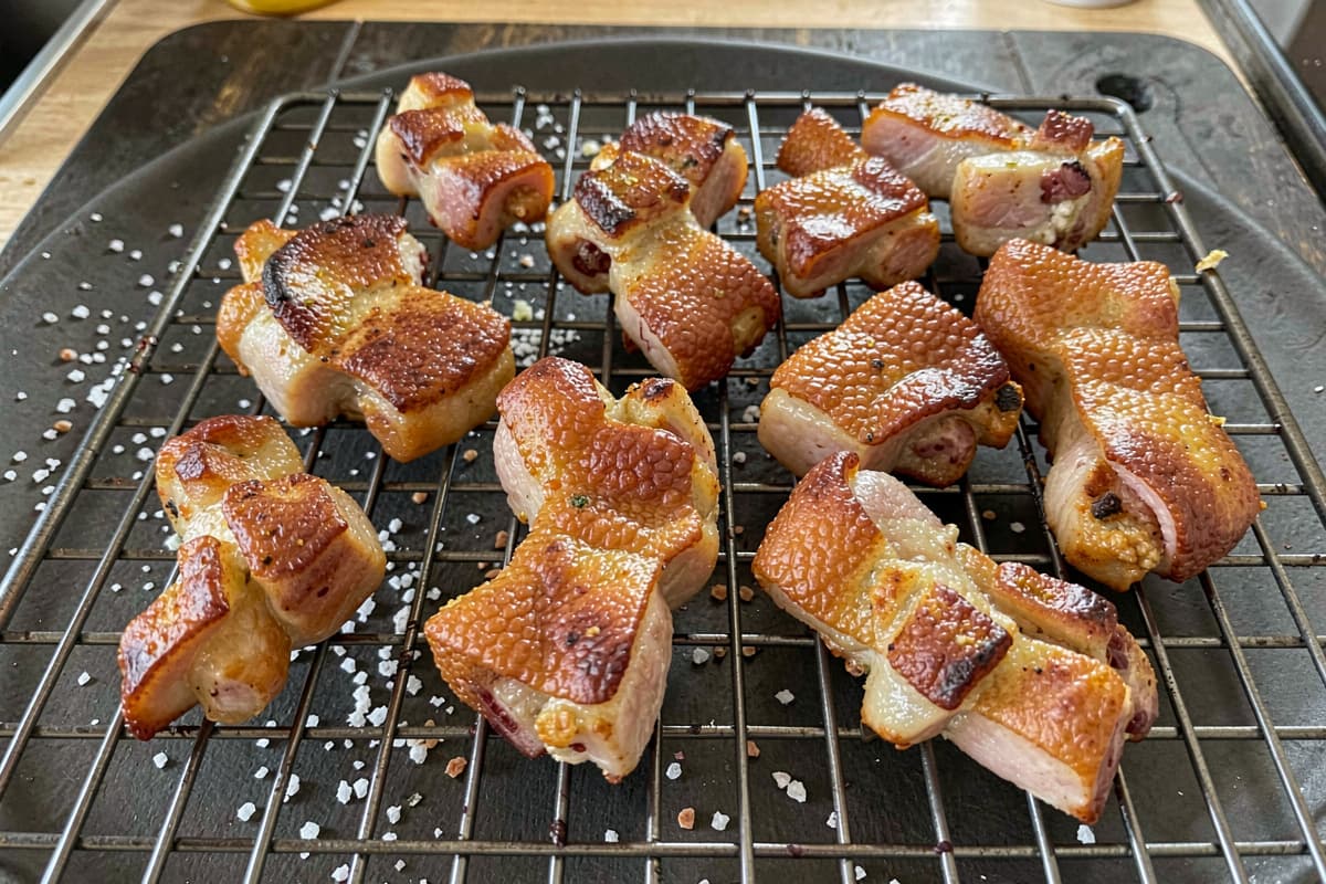 Pieces of pork crackling on a wire rack, golden-blistered surface, deeply puffed and shattering, scattered salt, dark cutting board background