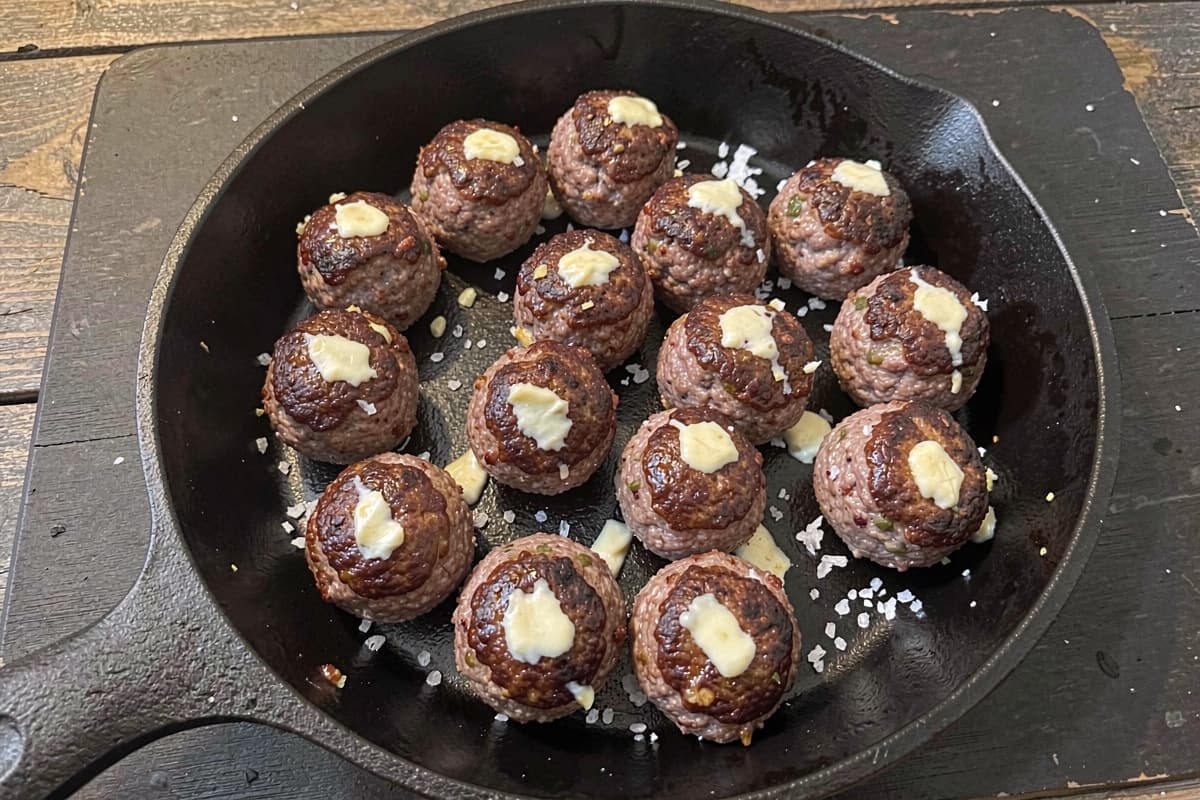 Sixteen pork meatballs in a cast iron skillet, deep brown crust on all sides, glistening with butter, scattered salt, dark wood underneath