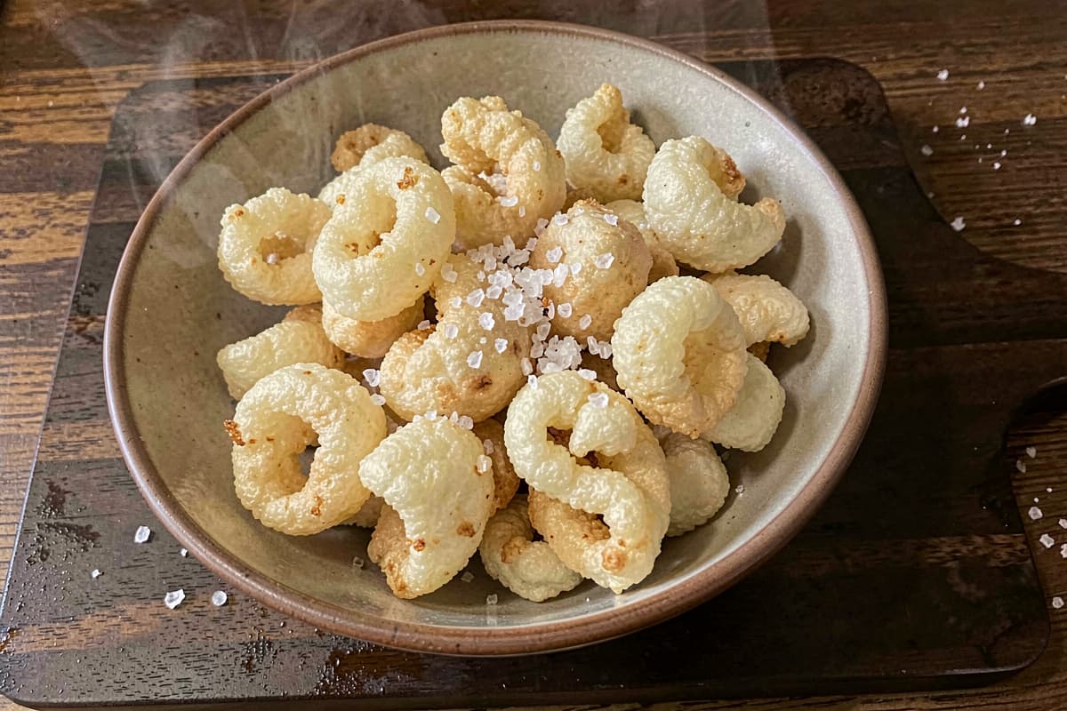 Bowl of homemade pork rinds, pale golden puffed crispy texture, scattered salt flakes, dark wooden background, freshly fried with steam rising slightly