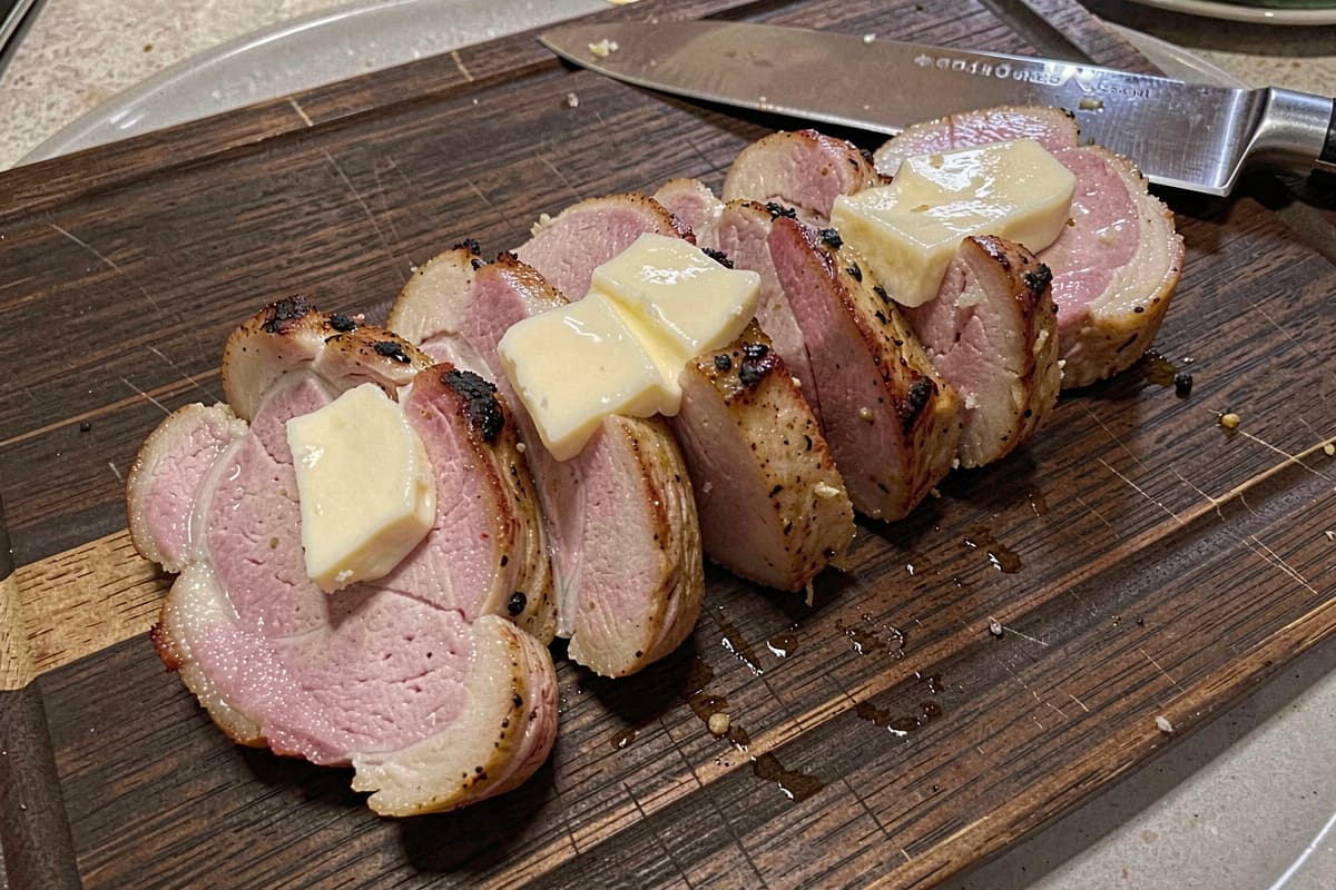 Sliced pork tenderloin on a wooden cutting board, golden seared exterior, slight pink interior, half-inch medallions fanned out, butter glistening on top