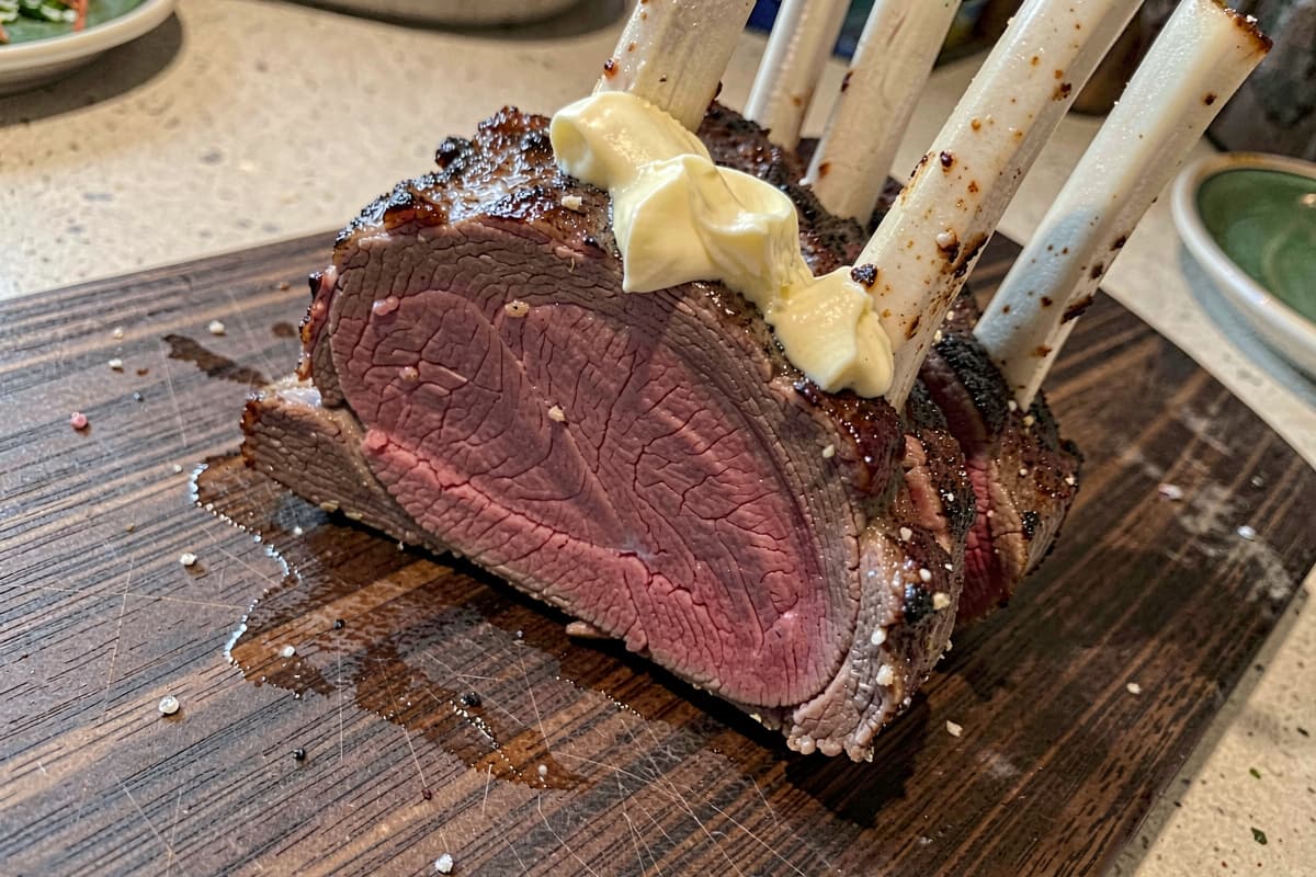 Frenched rack of lamb sliced between bones on a wooden cutting board, deep brown seared crust, rosy medium-rare interior, white bones standing up, butter glistening on the surface
