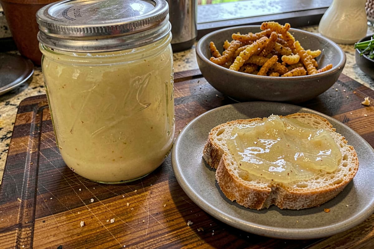 Glass mason jar of rendered schmaltz on a wooden counter, creamy pale yellow, golden gribenes in a small bowl beside it, slice of bread spread with schmaltz