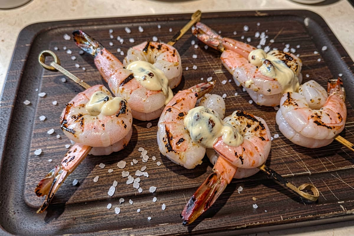 Four grilled shrimp skewers on a wooden cutting board, opaque pink shrimp with light grill marks, butter glistening on each, scattered salt, skewers fanned out at angles