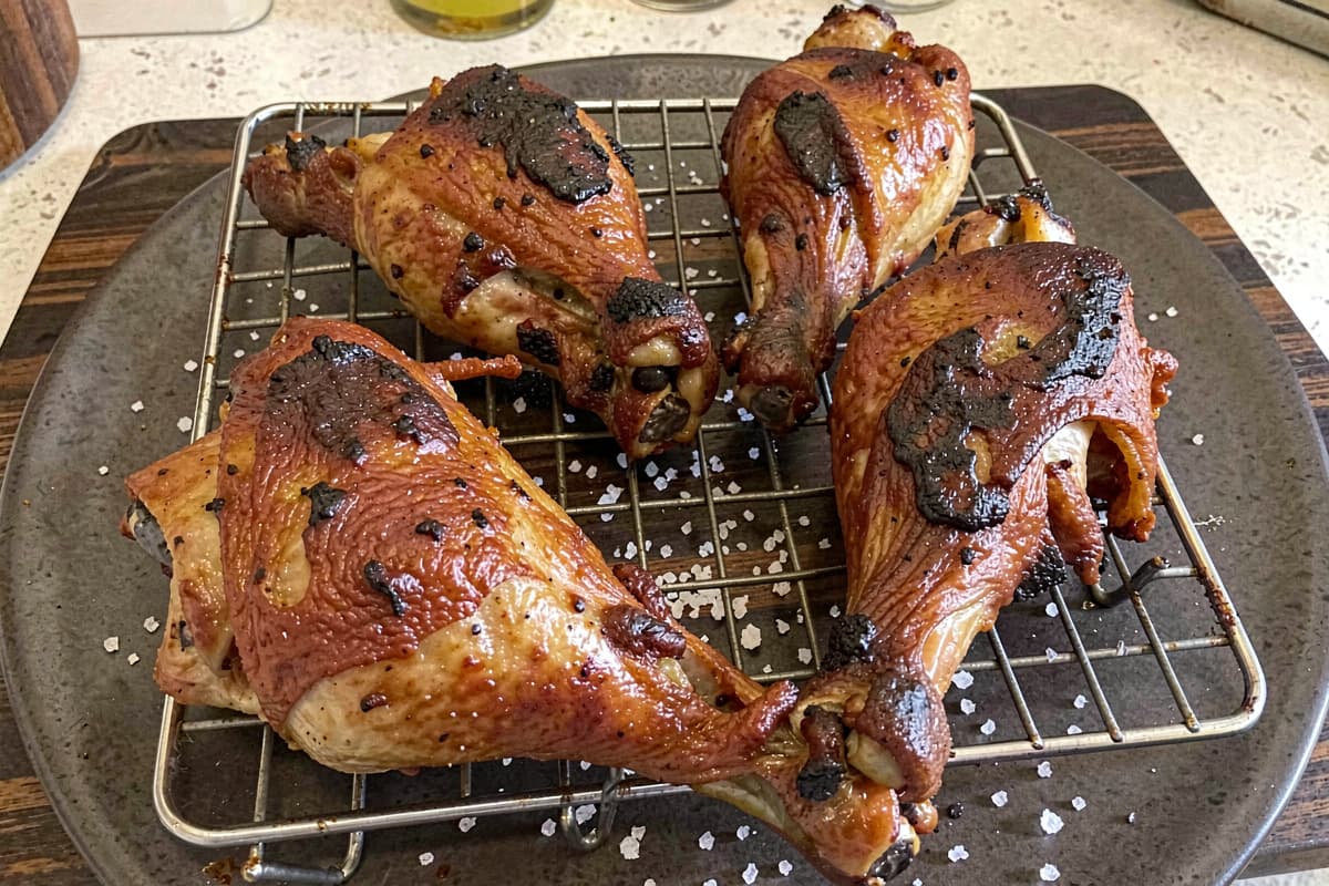 Four smoked chicken leg quarters on a wire rack with deep amber crispy skin, joints visible, scattered salt around them on dark wood