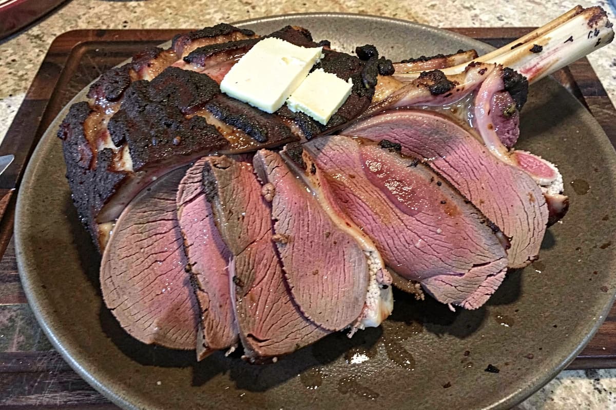 Sliced smoked leg of lamb on a wooden board, deep brown crust, rosy medium-rare interior, butter glistening on the surface, bone visible at one end