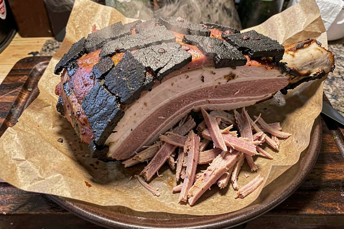 Whole smoked pork shoulder on butcher paper with thick mahogany bark, bone visible at one end, fork-pulled shreds spilling onto the paper