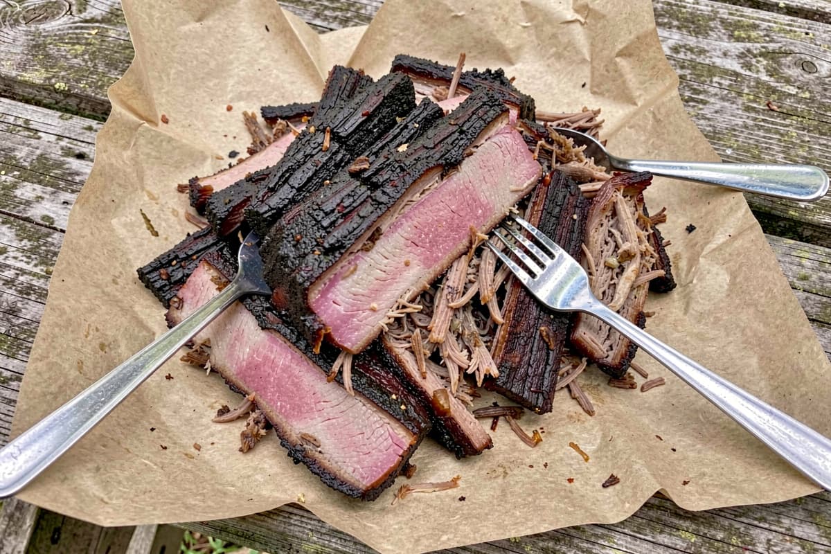 Pulled smoked pork shoulder on a wooden cutting board with two forks, dark bark visible on the outside, moist shredded interior