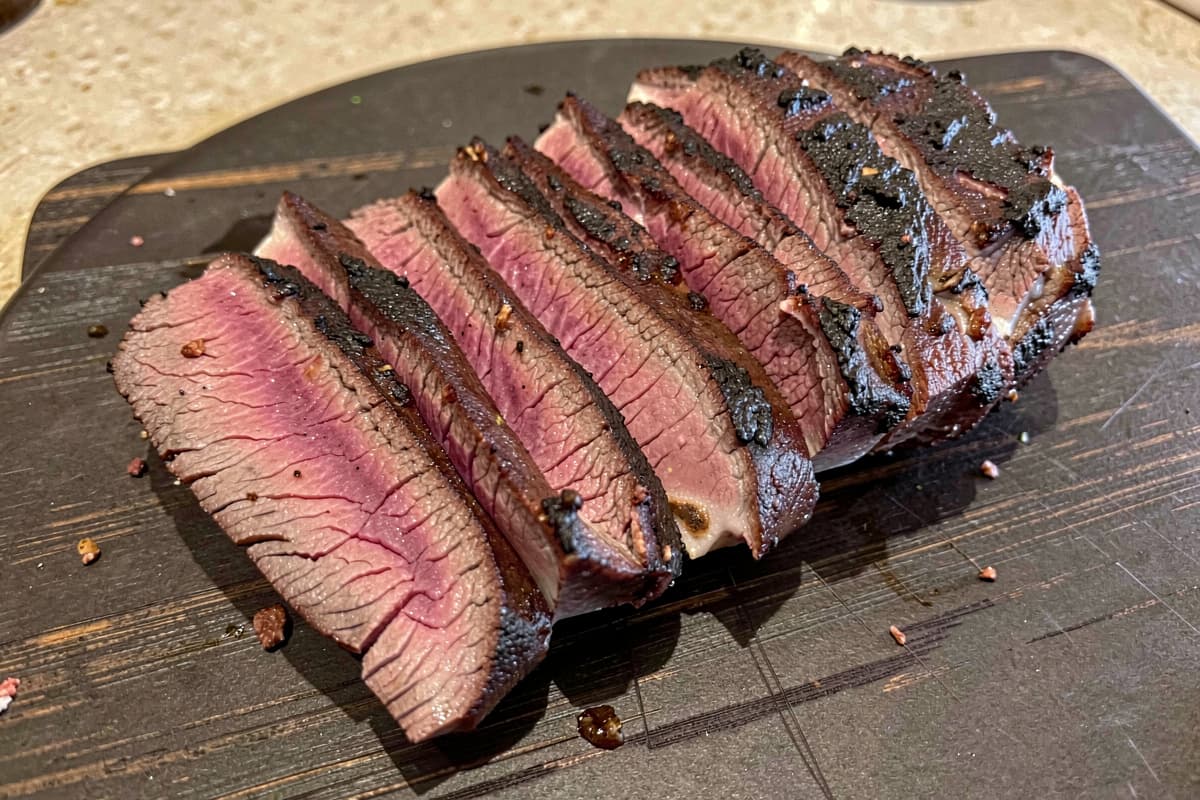 Sliced smoked tri-tip on a wooden cutting board, dark seared crust, pink smoke ring visible just under the surface, rosy medium-rare interior