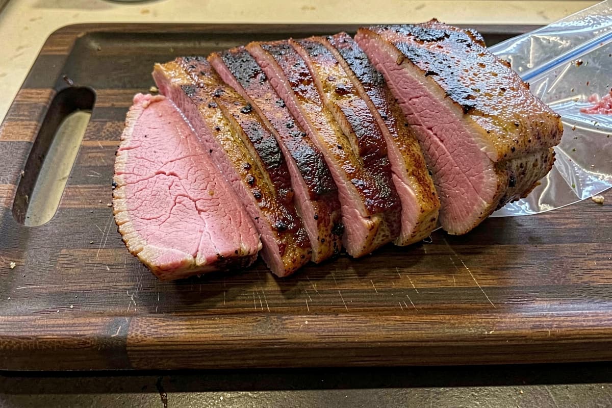 Sliced sous vide pork loin on a wooden cutting board, deep golden seared crust, edge-to-edge pink interior, half-inch slabs fanned out, sous vide bag visible on the side