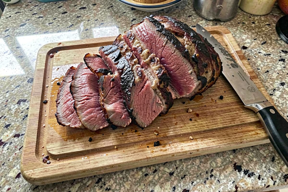 Sliced tri-tip roast on a wooden cutting board with a deep brown crust and pink medium-rare interior, butter melting on top