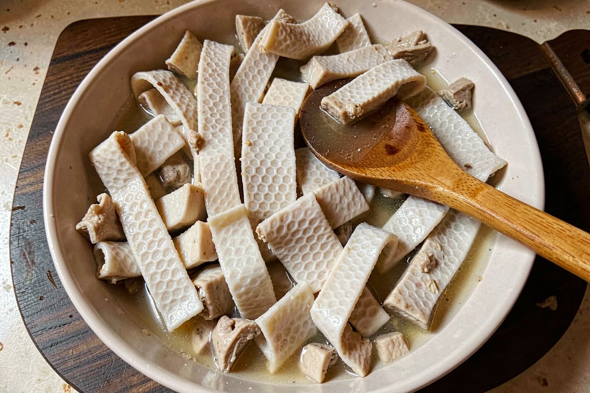Sliced cooked tripe ribbons in a wide bowl, white-pale honeycomb pattern visible, salty broth pooling around them, wooden spoon resting in the bowl
