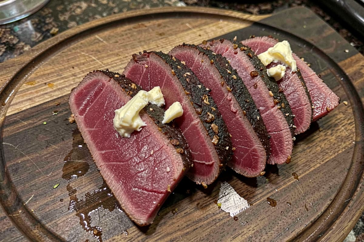 Sliced seared ahi tuna steak on a wooden cutting board, thin dark crusty exterior, deep red rare interior edge to edge, half-inch slabs fanned out, butter glistening