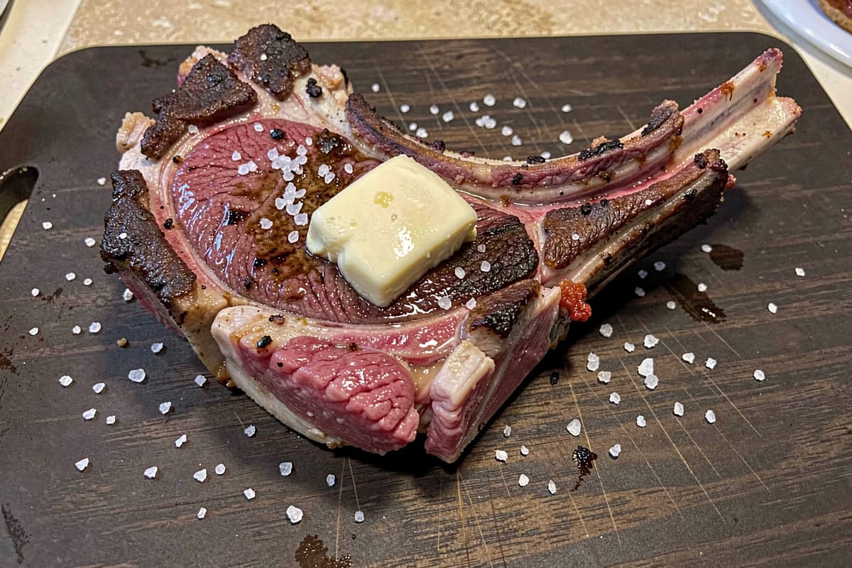 Single thick-cut veal rib chop on a wooden cutting board, deep brown seared crust, pale pink interior, rib bone protruding, butter glistening on top, scattered salt