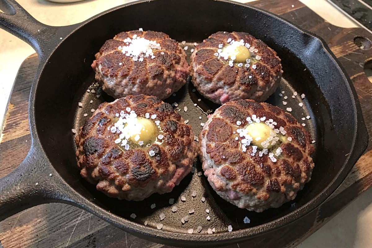 Four venison-pork burger patties in a cast iron skillet, deep brown crust, slight pink interior visible at the cut, scattered salt, glistening with butter